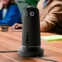 Owl Labs Meeting Owl 5 Pro 360 4K konferencijų kamera, pastatyta vertikaliai ant medinio stalo, su valdymo mygtukais ir apvaliu logotipu, fone išplaukusios žmonių figūros susitikimo aplinkoje.
