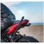 Artimas raudono motociklo vaizdas, stovinčio smėlingame paplūdimyje, su juodai apsirengusiu vairuotoju. Dėmesys skiriamas galinei daliai, įskaitant galinį žibintą ir dalį išmetimo sistemos, idealiai tinka stabiliai vaizdo filmav