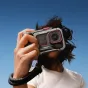 Insta360 Ace Pro 2 su Tilta Xplorer Grip Kit pilkos spalvos, ergonomiška rankena gatvės fotografijai ir miesto kelionėms, laikoma rankoje prieš aiškų mėlyną dangų