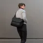 Peak Design Everyday Messenger V2 13L juodas nešiojamasis krepšys su paminkštinta petneša juosta, nešiojamas žmogaus prie pilkos sienos