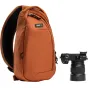 Think Tank TurnStyle 5L Urban Rust sling bag su juodu tinklo nugaros paneliu, su užtrauktuku pagrindiniu skyriumi, planšetinio kompiuterio kišene ir vandens butelio kišene, šalia juodos kameros su Sony objektyvu.