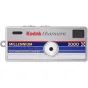 Kodak Charmera Millennium – sidabrinė raktų pakabuko skaitmeninė kamera su apvaliu objektyvu, mėlynos ir raudonos juostelės dizainu, tekstu 'MILLENNIUM' ir '2000', bei mažu juodu ieškikliu.