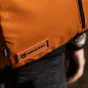 Artimas planas ryškiai oranžinio Wandrd PRVKE 21 Litro Sedona Orange Photo Bundle V3 su juodu užtrauktuku, šešiakampiu logotipu ir prekės ženklu, įspaustu ant odinės etiketės, dėvimas virš juodų kelnių, dešinėje pusėje matoma ne