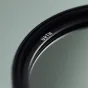 Artimas juodo URTH 55mm ND64 (6 žingsnių) objektyvo filtro vaizdas su sidabriniu įrašymu krašte, demonstruojantis stiklo paviršių ir sriegio detales neutralios pilkos fono atžvilgiu.