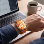 Tech-Protect oranžinė nailono apyrankė Huawei Watch Fit serijai, kvėpuojanti ir greitai pritvirtinama, dėvima ant riešo prie stalo su nešiojamuoju kompiuteriu ir kavos puodeliu.