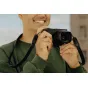 Peak Design Form Leather Long juodas odinis kameros diržas su žemaprofiliais greitais jungikliais, pritvirtintas prie Leica kameros, kurią laiko besišypsantis fotografas žaliame megztinyje.