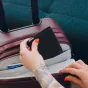 Peak Design Passport Wallet juodas kelioninis piniginė su magnetiniu užsegimu, dedama į bordo lagaminą, ranka su raudonu nagų laku, matoma National Airlines First Class kortelė.