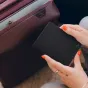 Juodas Peak Design Passport Wallet laikomas rankose su oranžiniu nagų laku, šalia bordo lagamino, akcentuojant jo ploną dizainą ir magnetinį užsegimą.